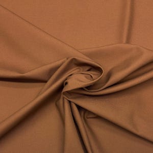 May include: Close-up of a swatch of fabric in a warm, earthy brown color. The fabric is smooth and appears to be a medium weight, with soft folds and subtle shadows. The texture suggests a natural fiber like cotton or linen.