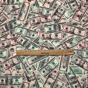 Pode incluir: Um close-up de um tecido com um padr&atilde;o de notas de d&oacute;lar americano em v&aacute;rias denomina&ccedil;&otilde;es. As notas s&atilde;o impressas em tons de verde, preto e branco, e est&atilde;o dispostas em um padr&atilde;o aleat&oacute;rio. Uma r&eacute;gua de madeira &eacute; colocada sobre o tecido, com o texto "WESTCOTT" vis&iacute;vel.