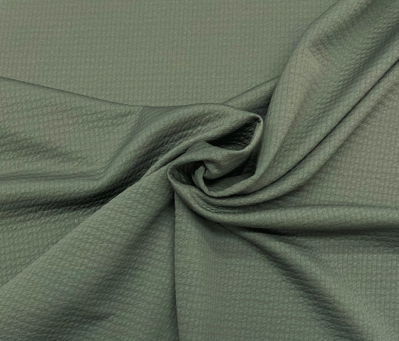 May include: Close-up of a folded piece of olive green fabric. The fabric has a textured, quilted appearance with a subtle sheen. The folds create shadows and highlights, emphasizing the fabric's texture and color. The material appears soft and pliable.