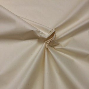 May include: A close-up of a cream-colored fabric with a subtle speckled texture. The fabric is folded and draped, showcasing its soft and smooth surface.