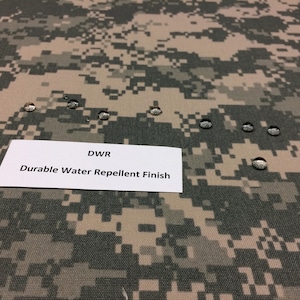 以下が含まれることがあります： 表面に水滴がついた迷彩柄の生地のクローズアップ。画像の中央には、「DWR Durable Water Repellent Finish」というテキストが書かれた白いラベルが見える。