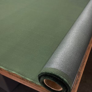 May include: A roll of dark green fabric with a silver sheen. The fabric is rolled up on a wooden surface.