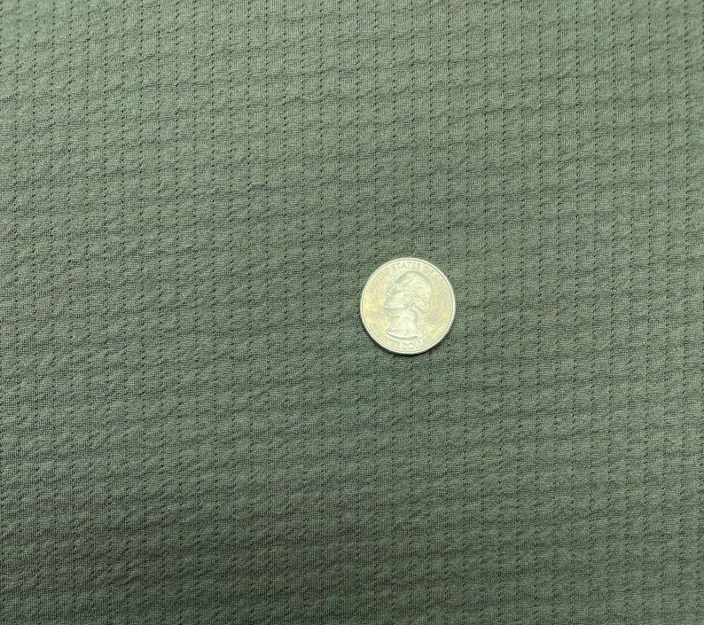 May include: Close-up of olive green fabric with a textured, waffle-like pattern. A US quarter coin is placed on the fabric for size comparison. The fabric appears to be a knit or woven material.