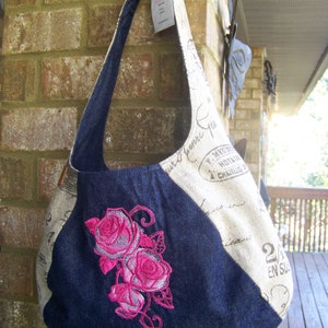 Könnte beinhalten: Eine blaue Jeans- und beige Stofftasche mit einem rosa gestickten Blumendesign. Die Tasche hat einen langen Riemen und ist mit französischem Text verziert.