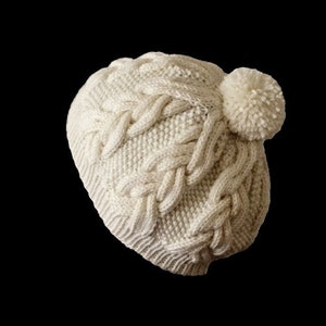 May include: A white knitted beanie with a large pom pom on top. The beanie has a cable knit pattern.