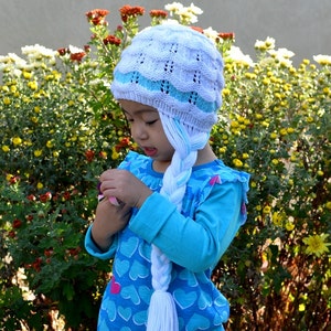May include: A young girl wearing a white and blue knitted hat with a long braid attached. The hat has a blue stripe and the braid is white. She is wearing a blue shirt with white hearts and is standing in front of a bush of yellow flowers.