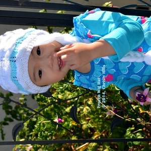 May include: A young child wearing a white and blue knitted hat with a long white braid. The hat has a blue stripe and the braid is attached to the hat. The child is wearing a blue shirt with white hearts. The text "Frozen Wig Hat &copy; 2014, Christy Hills" is visible in the image.