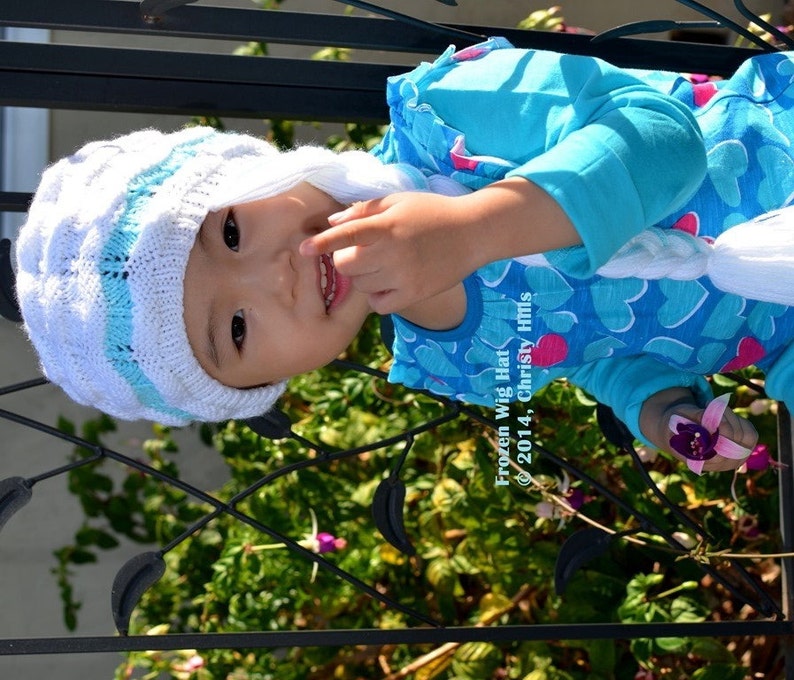 May include: A young child wearing a white and blue knitted hat with a long white braid. The hat has a blue stripe and the braid is attached to the hat. The child is wearing a blue shirt with white hearts. The text "Frozen Wig Hat &copy; 2014, Christy Hills" is visible in the image.