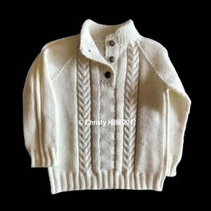 Könnte beinhalten: Ein weißer Strickpullover mit Stehkragen und Zopfmuster. Der Pullover hat einen Knopfverschluss am Hals.