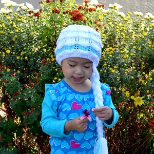 May include: A young girl wearing a blue and white knit hat with a long white braid attached. She is wearing a blue shirt with pink hearts and is holding a purple flower.