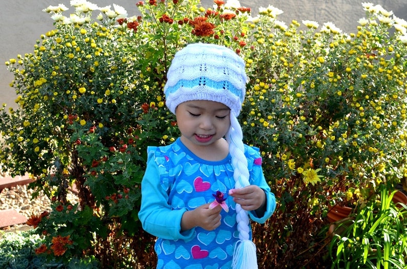 May include: A young girl wearing a blue and white knit hat with a long white braid attached. She is wearing a blue shirt with pink hearts and is holding a purple flower.