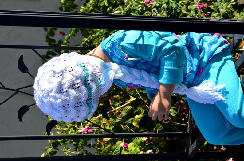 May include: A young person wearing a white knitted hat with a blue and white braid attached. The hat has a white and blue pattern. The person is wearing a blue shirt with a white and blue pattern. The person is sitting on a black metal railing.
