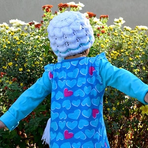 May include: A young child wearing a blue shirt with a heart pattern and a white crocheted hat stands in front of a bush of yellow flowers.