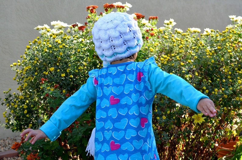 May include: A young child wearing a blue shirt with a heart pattern and a white crocheted hat stands in front of a bush of yellow flowers.