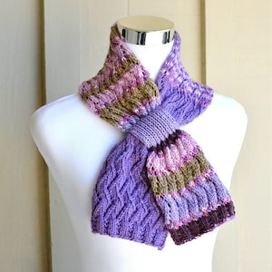 Peut inclure: Une écharpe en tricot de couleur violette, rose et marron avec un motif en tricot torsadé. L'écharpe est nouée et présentée sur un mannequin blanc.