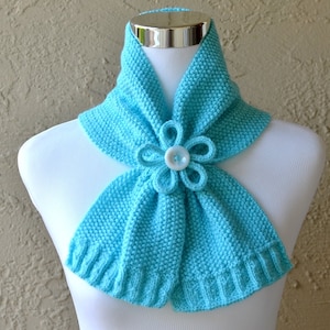 May include: A turquoise blue knitted scarf with a white button flower accent.