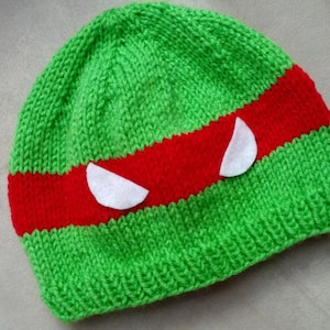 May include: Green knitted hat with a red stripe and white felt eyes. The hat is designed to look like a Teenage Mutant Ninja Turtle.
