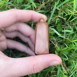 Könnte beinhalten: Ein rechteckiger Anhänger mit einer kupferfarbenen Drahtschlaufe. Der Anhänger hat eine einfarbige, hellbraune Farbe mit glänzendem Finish. Der Hintergrund ist grünes Gras.