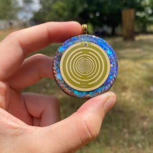 May include: Round pendant with a gold-colored bail. The pendant has a blue and iridescent border surrounding a light green center with a spiral design. The text "MWD EMFs" is visible on the pendant.