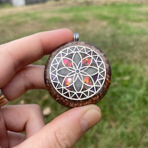 Könnte beinhalten: Runder Anhänger mit einem silbernen geometrischen Design auf einem braunen und rot gesprenkelten Hintergrund. Der Anhänger hat eine silberne Öse für eine Kette. Die Mitte des Anhängers hat ein Blumendesign mit roten und orangen Akzenten.