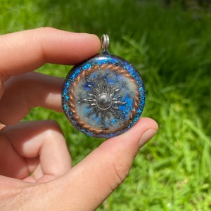 Könnte beinhalten: Runder Anhänger mit einem silbernen Sonnendesign in der Mitte. Der Anhänger hat einen blau-silbernen Hintergrund mit einem kupferfarbenen Rand. Er wird von einer silbernen Schlaufe gehalten.