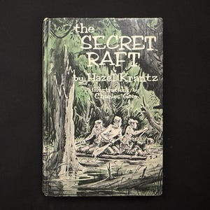 Puede incluir: Portada de libro vintage de "The Secret Raft" de Hazel Krantz, con ilustraciones de Charles Geer. La portada presenta una ilustración en verde y blanco de tres figuras en una balsa en una zona pantanosa, con el título en letras blancas grandes.