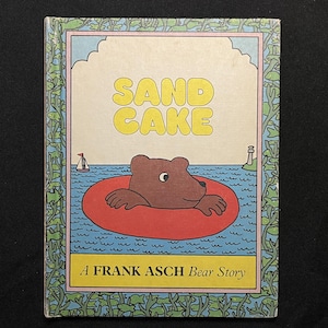 May include: A book cover with a yellow and orange title "SAND CAKE" and a brown bear wearing a red inner tube floating in blue water. The book is titled "A FRANK ASCH Bear Story".