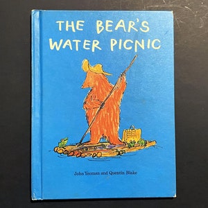 以下が含まれることがあります： 青い表紙の絵本「The Bear's Water Picnic」。表紙には、長い棒を持ったオレンジ色のクマが筏に乗っているイラストが描かれています。著者の名前は下部に印刷されています。