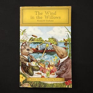 Op de afbeelding: Een vintage exemplaar van "De Wind in de Wilgen" van Kenneth Grahame. De omslag toont een illustratie van antropomorfe dieren die picknicken bij een rivier. De titel staat in een gele banner, met "Junior Classics for Young Readers" onderaan.