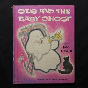 Puede incluir: Una portada de libro rosa con el título "Gus and the Baby Ghost" en letras blancas. La portada presenta una ilustración de un fantasma sentado en una mecedora, leyendo un libro. Un gato negro se sienta en el suelo a la derecha de la mecedora.