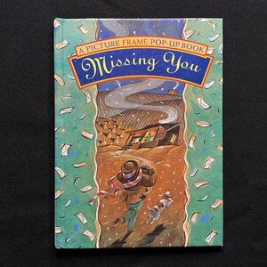 Puede incluir: Un libro desplegable titulado "Missing You" con un diseño de marco de imagen. La portada presenta a un hombre con sombrero y bufanda caminando con un perro, rodeado de letras que caen.