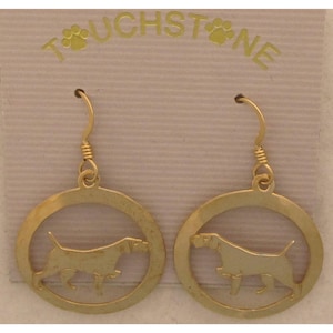 May include: Gold-colored dog earrings. Each earring features a circular frame with a cutout silhouette of a dog. The earrings hang from a curved hook. The word "TOUCHSTONE" is printed at the top.