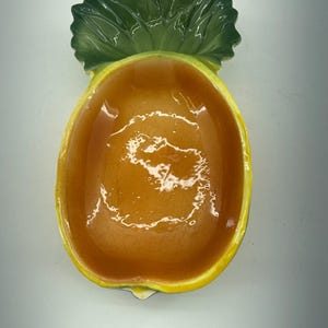 May include: A ceramic pineapple-shaped dish with a yellow and orange interior. The dish has a green leafy top and a yellow rim. The interior is a glossy orange color. This decorative dish is perfect for serving snacks or as a kitchen accent.