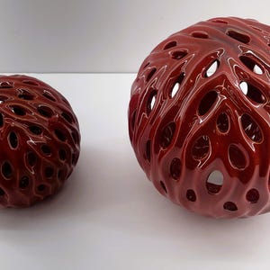 Puede incluir: Dos esferas decorativas burdeos brillantes con intrincados diseños calados. Las esferas varían en tamaño, una es más grande que la otra. Las superficies tienen un patrón fluido y orgánico, creando una textura visual única. Objetos decorativos para la decoración del hogar.