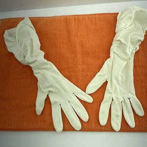 Peut inclure: Une paire de longs gants d'opéra ivoire. Les gants sont faits d'un tissu lisse et présentent un design froncé sur le haut du bras. Les gants sont posés sur une surface orange texturée.