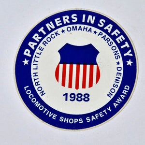 May include: A blue and white circular sticker with the text "Partners in Safety" and "Locomotive Shops Safety Award". The sticker also features a shield with red and white stripes and the year "1988".