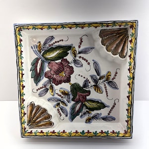 Peut inclure: Plat carré en céramique avec un fond blanc et un motif floral dans les tons de vert, bleu, violet et rouge. Le plat a une bordure jaune et rouge et trois sections surélevées.