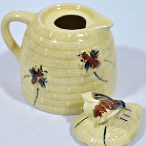 May include: A yellow ceramic creamer with a beehive design and a matching lid. The creamer features a handle and is decorated with painted bees. The lid is shaped like a honeycomb with a bee on top. A charming addition to any kitchen.