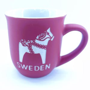May include: A magenta ceramic mug with a white Dala horse design and the word "SWEDEN" printed on the front. The mug has a white interior and a curved handle.