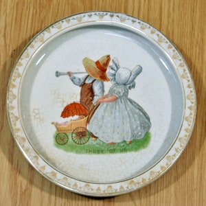 Puede incluir: Un plato de cerámica blanco con un borde dorado y una escena pintada de dos niños empujando un cochecito. Los niños llevan sombreros y la niña lleva un vestido azul con lunares blancos. El texto "THREE OF US" está impreso en el plato.