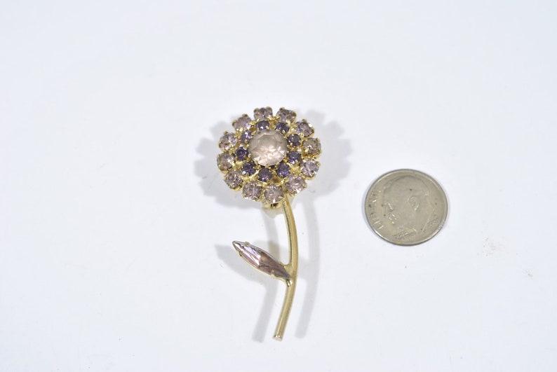 May include: A gold-toned flower brooch with a clear center stone and purple rhinestones. The flower has a stem and a leaf.