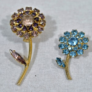 May include: Two gold tone flower brooches with rhinestone accents. One brooch features a purple and pink flower with a clear center stone. The other brooch features a blue flower with a light blue center stone.