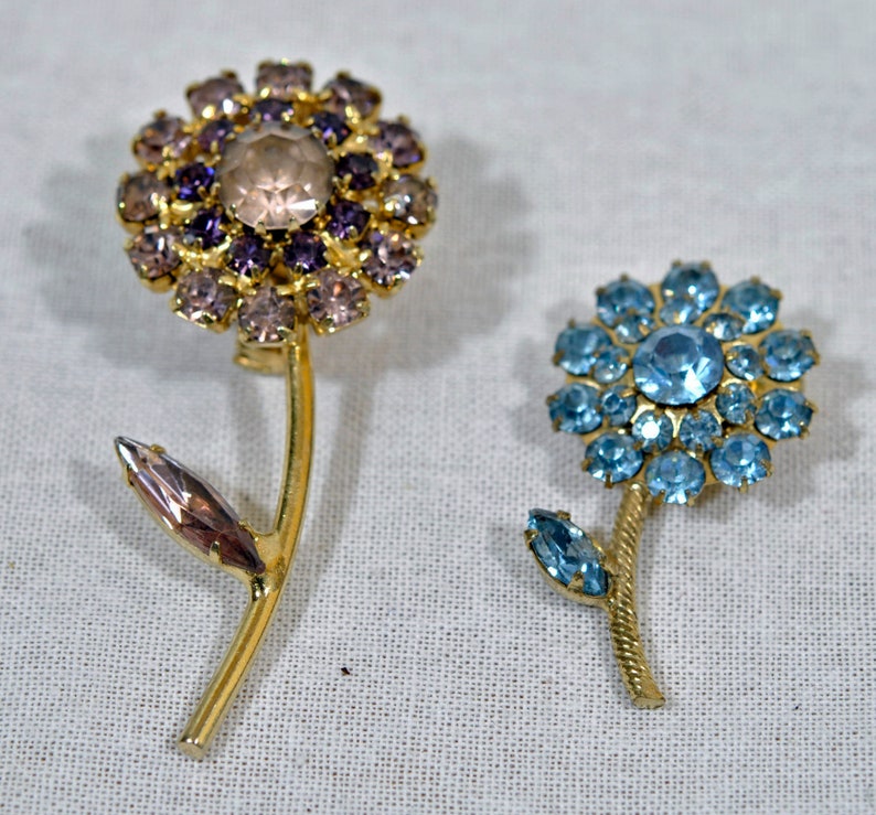 May include: Two gold tone flower brooches with rhinestone accents. One brooch features a purple and pink flower with a clear center stone. The other brooch features a blue flower with a light blue center stone.