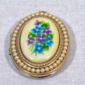 May include: A gold locket with a pearl border and a white oval cameo featuring a painted floral design of purple and blue flowers.