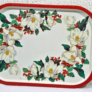 Könnte beinhalten: Ein weißes Serviertablett aus Metall mit rotem Rand. Das Tablett ist mit einem floralen Design aus weißen Magnolienblüten, grünen Blättern und roten Beeren verziert.