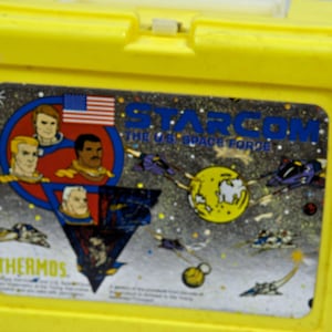 Könnte beinhalten: Gelbe Plastik-Lunchbox mit einem Starcom: The US Space Force Logo. Die Lunchbox zeigt ein Cartoon-Bild einer Weltraumszene mit Astronauten und Raumschiffen. Die Lunchbox ist von Thermos.