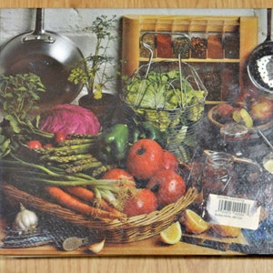 May include: A still life image of a variety of fresh vegetables, including red cabbage, green peppers, asparagus, carrots, and onions. The vegetables are arranged in a wicker basket with a white garlic bulb and a few green sprigs of herbs. There are also jars of preserved food and a metal colander filled with lettuce in the background.