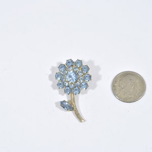 May include: A gold-toned flower brooch with a light blue rhinestone center and light blue rhinestones around the edges. The flower has a stem and a leaf.