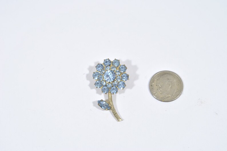 May include: A gold-toned flower brooch with a light blue rhinestone center and light blue rhinestones around the edges. The flower has a stem and a leaf.