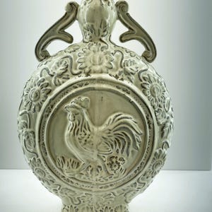 Pode incluir: Vaso de cerâmica branco sujo com relevo de galo. O vaso tem um corpo redondo com detalhes florais e de volutas, duas alças curvas e uma base quadrada. O galo está centrado em uma moldura circular.